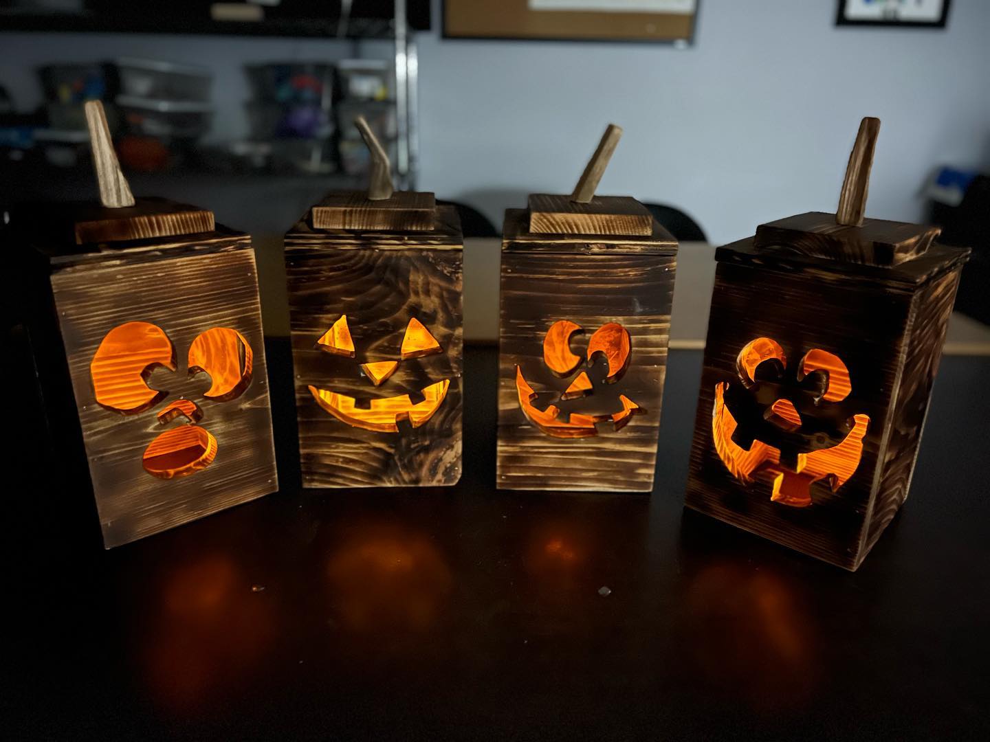 Four rustic wooden jack-o'-lantern lanterns with glowing orange lights, arranged in a row on a dark table.