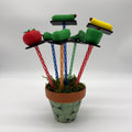 Miniature plastic vegetable stakes including a red tomato, green cucumber, and a yellow squash, mounted on colorful twisted sticks in a patterned green pot with faux moss.