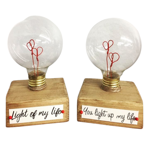 Two decorative light bulbs with clear glass globes, red heart-shaped filaments, and square wooden bases. The left base reads 'Light of my life' and the right base reads 'You light up my life' in black script with small red heart accents.