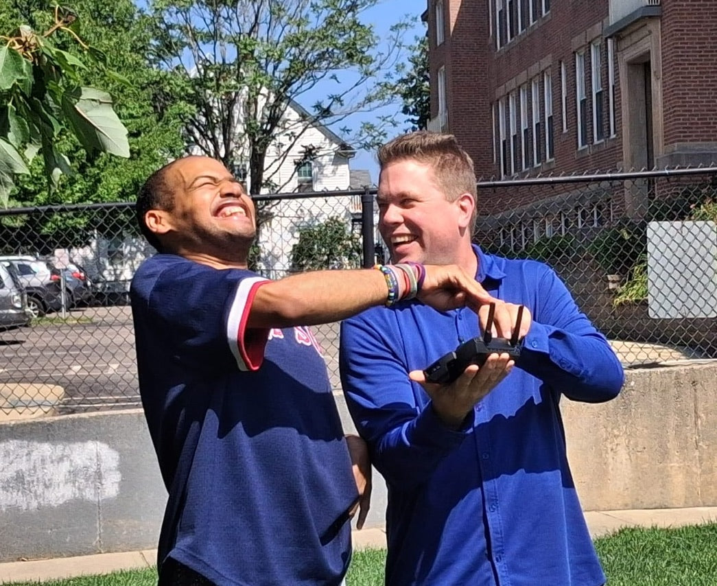 Two men in blue shirts interacting with a controller outdoors with a building and trees in the background