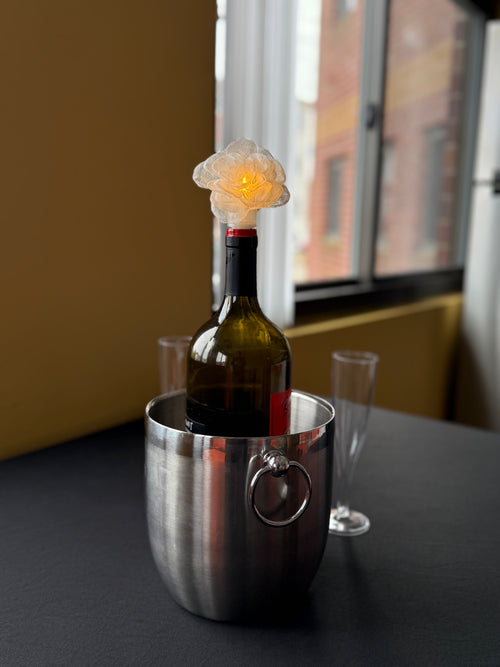 Wine bottle in a metal cooler with a decorative rose light on top, set against a windowed wall.