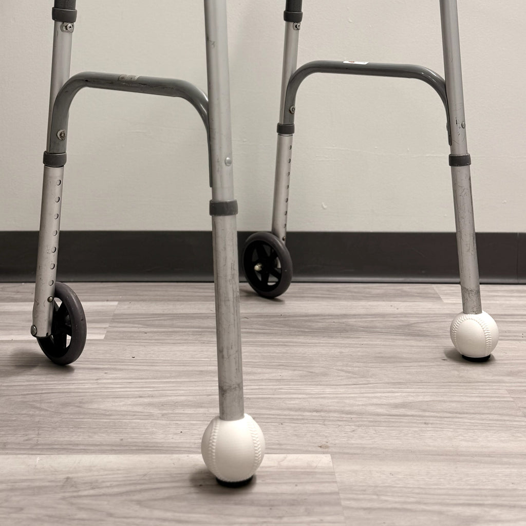 A silver metal walker with black handles, black wheels, and white ball castors at the bottom, standing on a light wood floor against a plain wall.