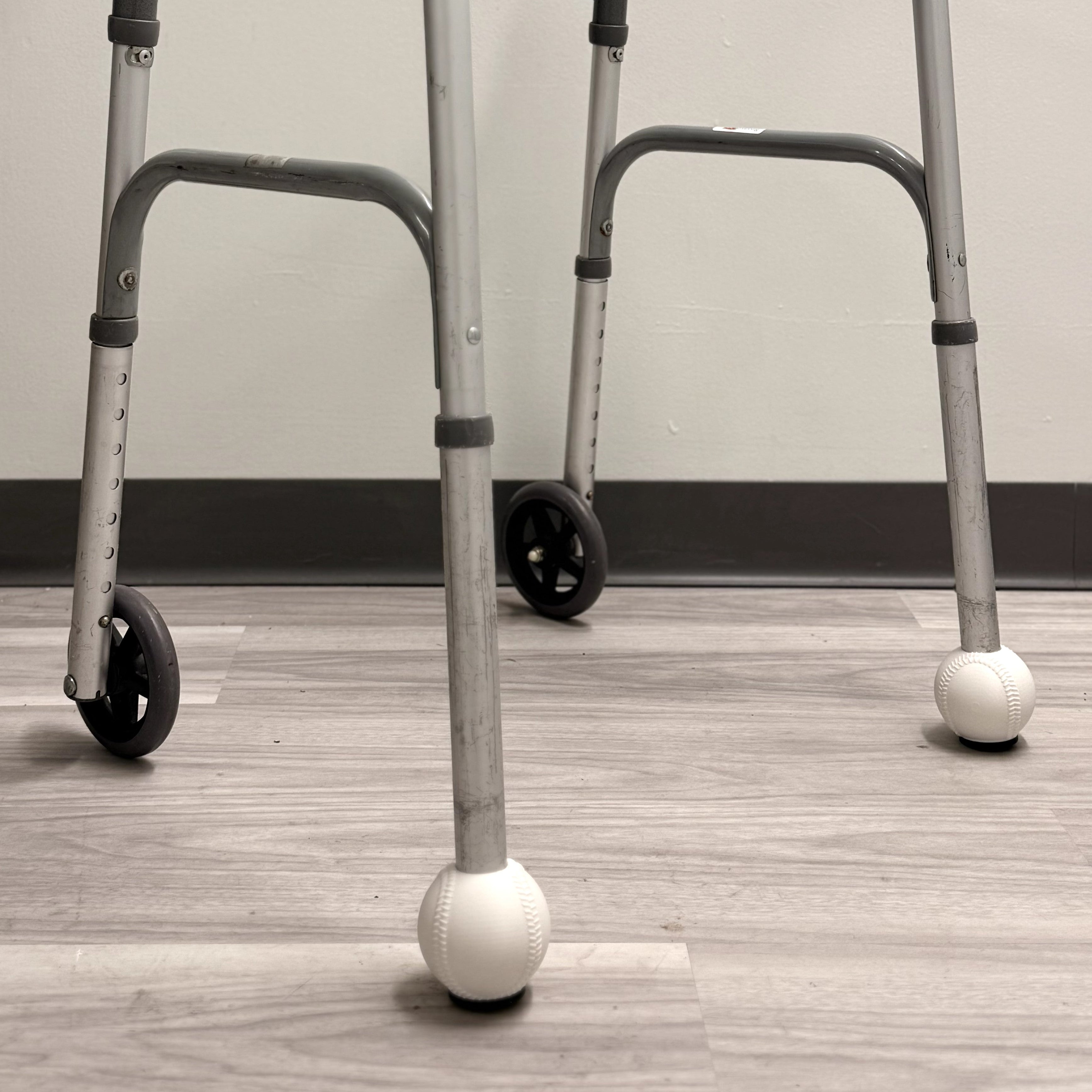 A silver metal walker with black handles, black wheels, and white ball castors at the bottom, standing on a light wood floor against a plain wall.